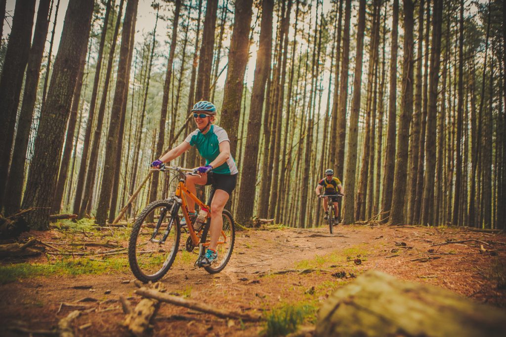 Female mountain biker riding through the forest with a fellow cycling a few yards behind.