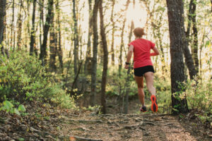 White woman with brown hair, dressed in red shirt, blacks shorts, and red shoes runs on a mountain trail in the daytime.