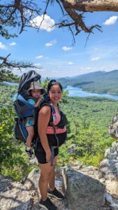 Pregnant Asian woman with a toddler in a hiking pack on her back smiles at the camera with a beautiful sunny mountain vista behind her.