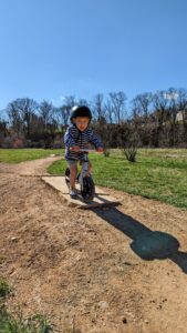 Toddler boy riding a bike on a dirt path wearing a black helmet and a jacket smiles at the camera on a sunny day.