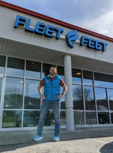 Man wearing a puffy blue vest over a short-sleeved shirt smiles at the camera. He has his hands on his hips, and he's standing in front of Fleet Feet, a running store.