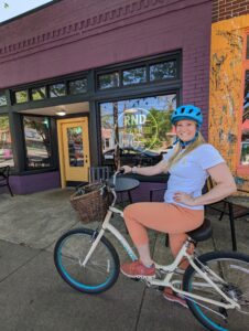 Blond woman on a bike wearing a blue helmet, white shirt, and orange pants poses in front of a coffee shop and smiles.