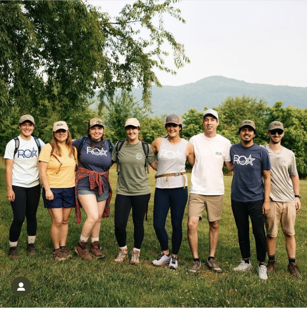 Group of eight white people posing and smiling for the camera outdoors with the mountains in the background.