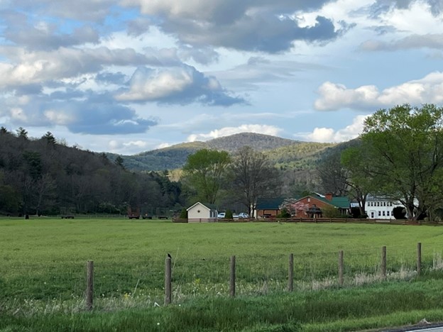Big green field with farms buildings in the background and the Blue Ridge Mountains behind that. The sky is blue with white fluffy clouds in the sky.