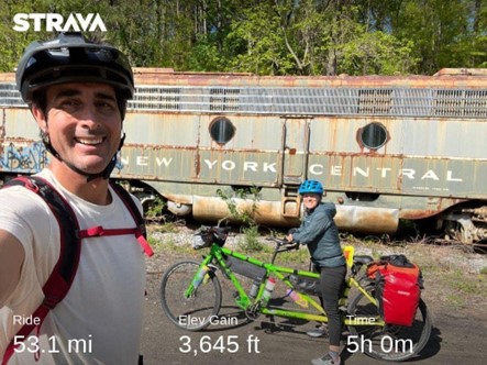 Man wearing a bike helmet white shirt, and a backpack takes a selfie with woman on a tandem bike and an old abandoned train car in the background.