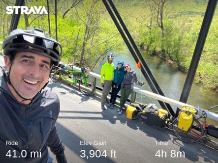 Man wearing a black zip-up jacket and bike helmet takes a selfie with three other cyclists posed and smiling in the background. There two tandem bikes are outfitted for a long trip, and they're on a bridge crossing over a wide creek on a sunny day.