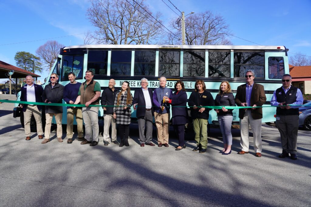 Several people pose for a ribbon cutting photo in front of a teal 20-passenger bus. 