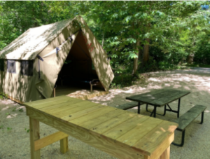 Campsite in a wooded area with a large canvas tent, tall wooden table and picnic table.